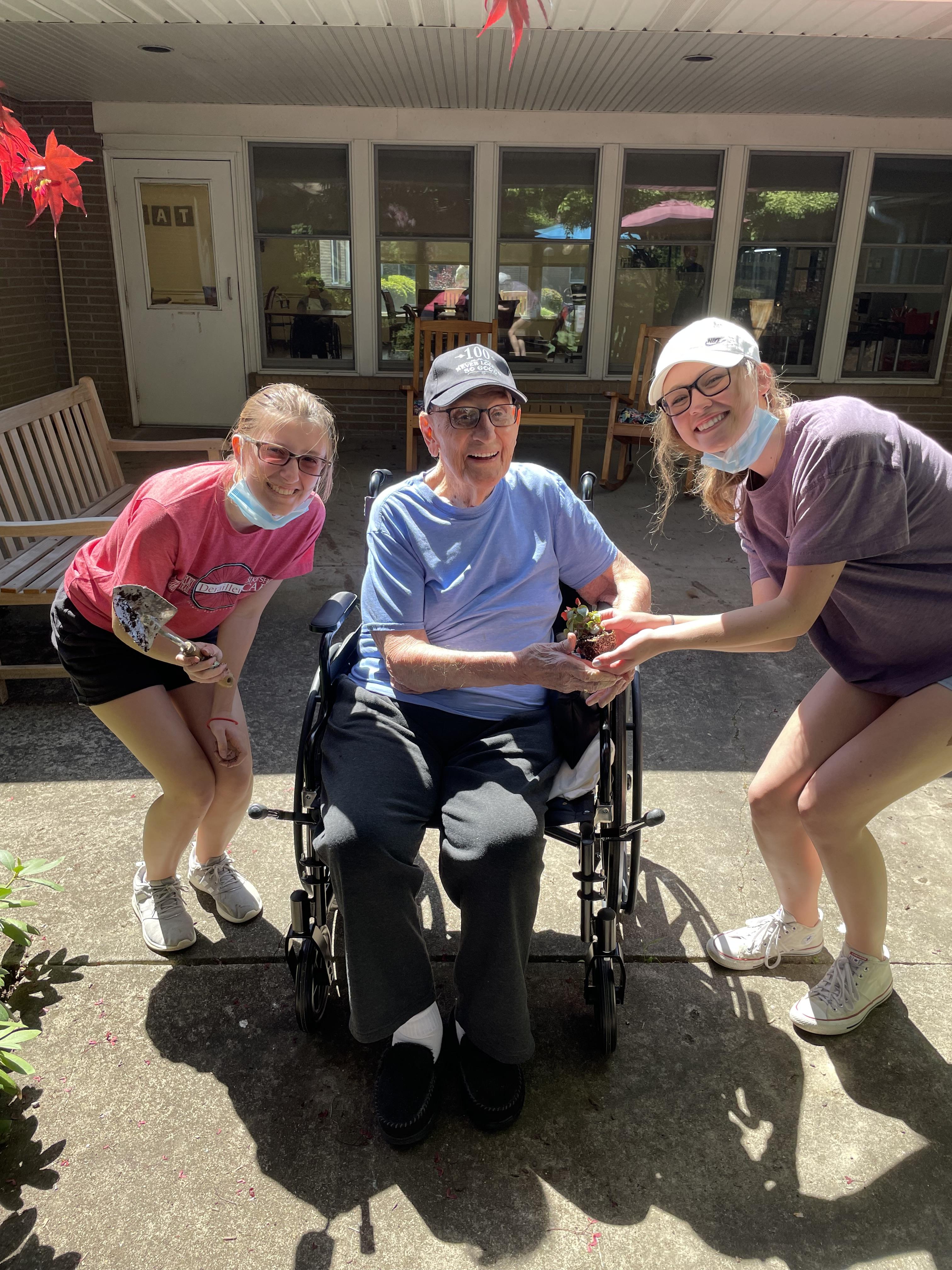pic of elderly resident in a wheelchair with 2 students outside of a facility